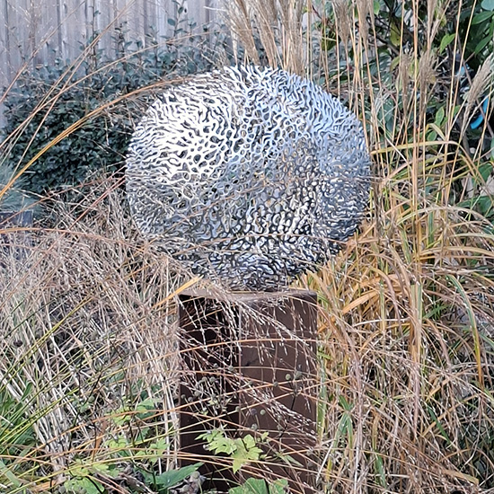 Stalen beeld met organische uitsparingen op een sokkel, geplaatst in het riet bij de waterkant.