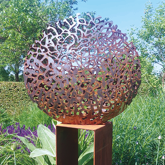 Rondvormige cortenstalen beeld met organische uitsparingen die doen denken aan vormen uit de zee, geplaatst op een sokkel buiten tussen de planten.