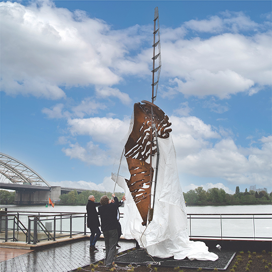 Twee mensen trekken het doek van een cortenstalen beeld tijdens de onthulling op de kade.