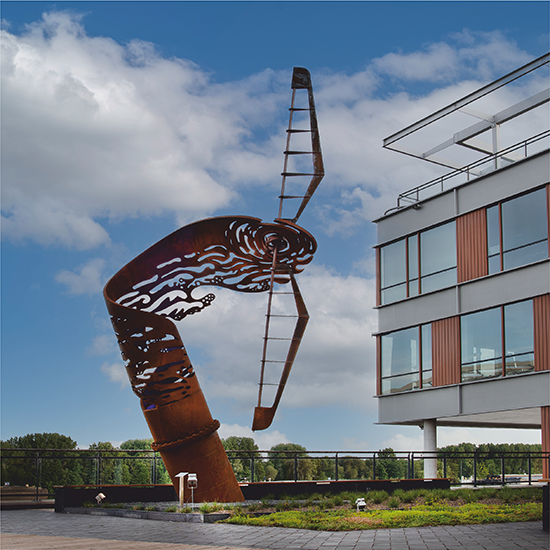 Groot cortenstalen beeld met nautisch geïnspireerde vormen, geplaatst buiten naast een gebouw.