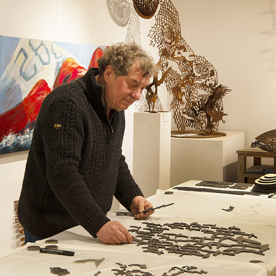 Kunstenaar Marco Käller werkt aan een stalen beeld met vogelvormen in een galerie met cortenstalen beelden en een groot olieverfschilderij.